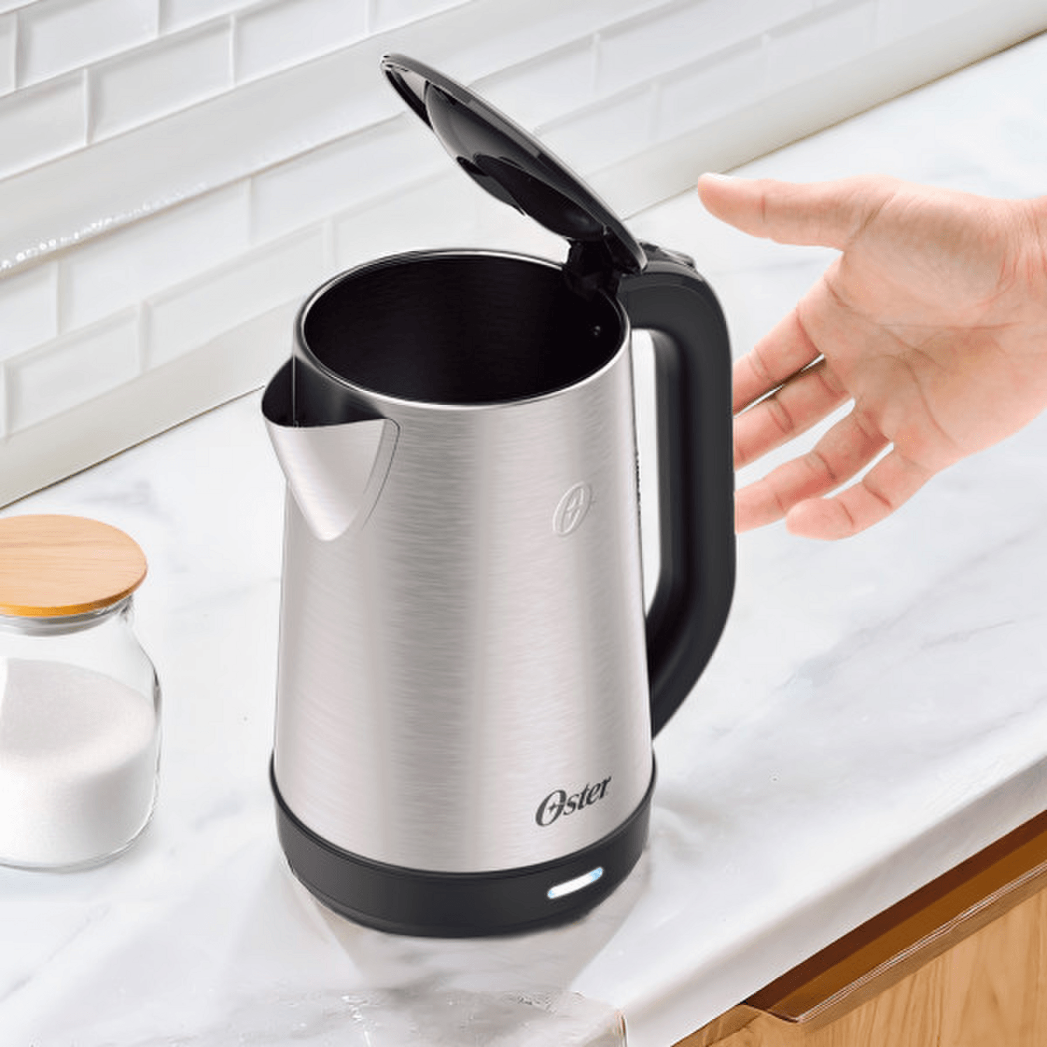 Hervidor Oster 2L 1200W Acero Inox Apagado Auto 2220516 en cocina sobre la mesa con mano cerca